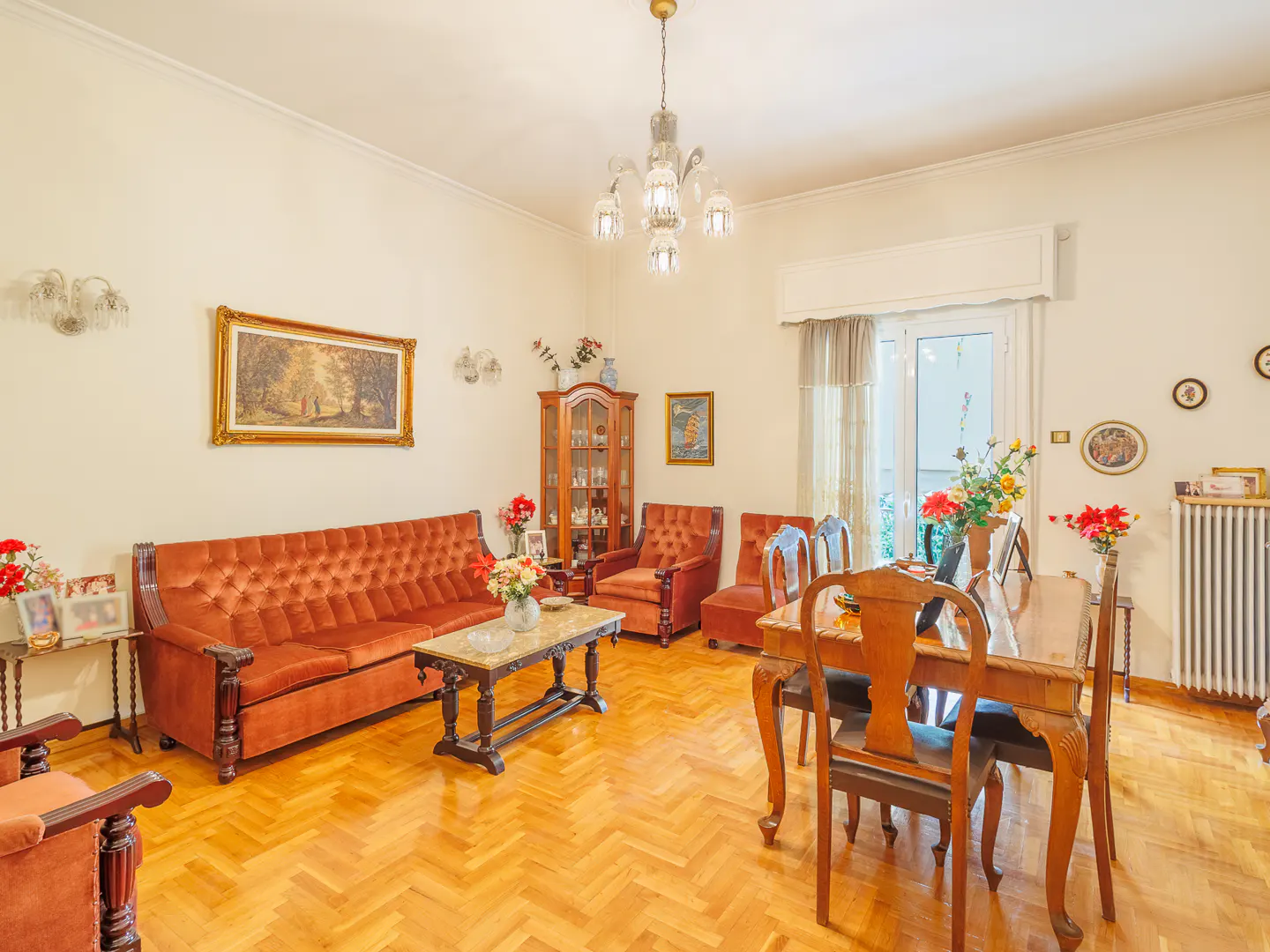 Classic living room with a tufted orange sofa, wooden dining set, parquet floors, and a crystal chandelier.