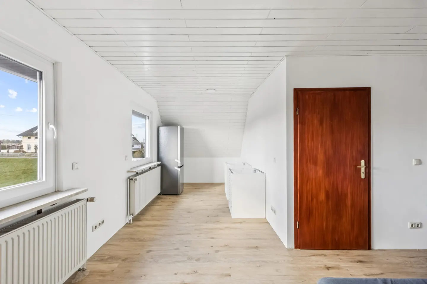 Bright attic room with white walls, wood floor, and a white plank ceiling. A brown door is on the right, and a silver refrigerator is on the left.