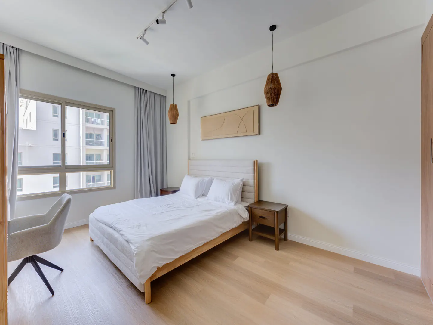Bright bedroom with a white bed, wooden frame, and white linens. A gray chair sits near a window with gray curtains. Wooden floors and nightstands.
