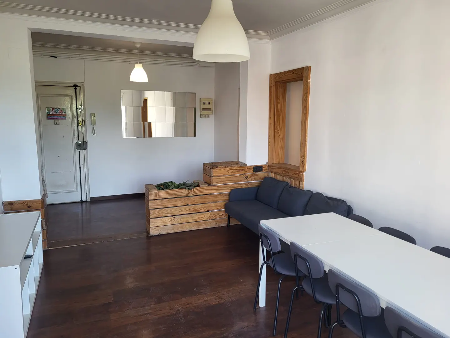 A bright living room with a white table, gray chairs, and a blue sofa. Wooden crates are used as decor. A white door is visible in the background.