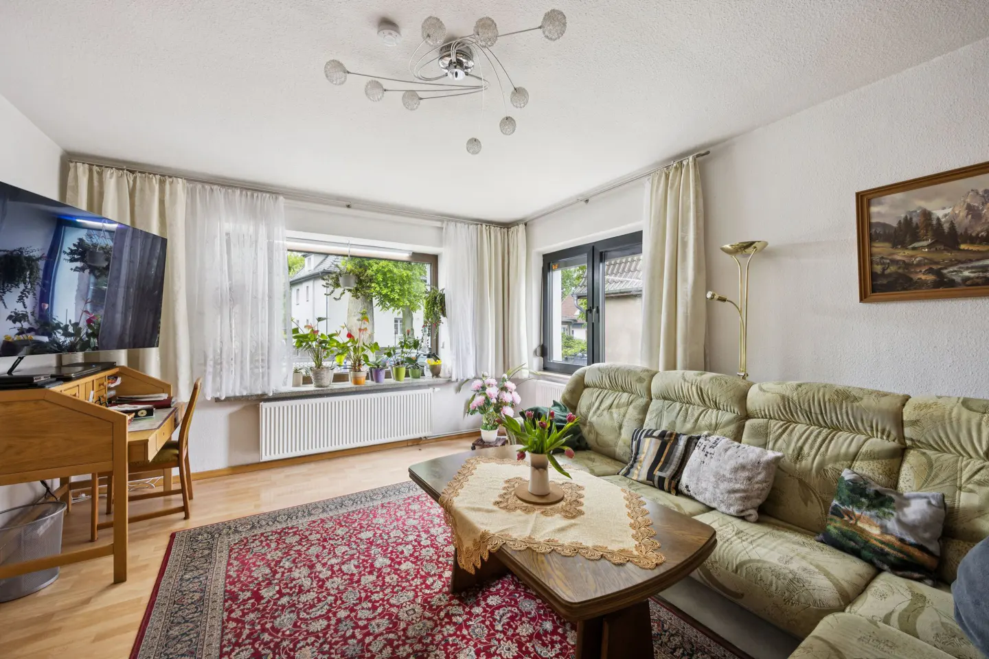 Living room with a green sofa, wooden table with flowers, and a red patterned rug. A desk and TV are on the left, with a window view of greenery.