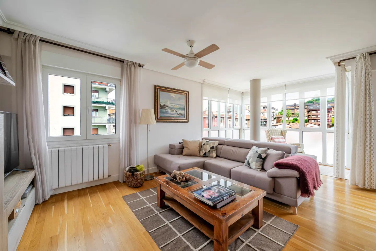 Bright living room with light wood floors, a grey sectional sofa, wooden coffee table, and large windows with city views.