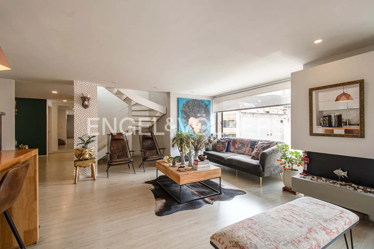 Bright, open living room with a gray sofa, wood coffee table, and two leather chairs. A white spiral staircase is visible in the background.