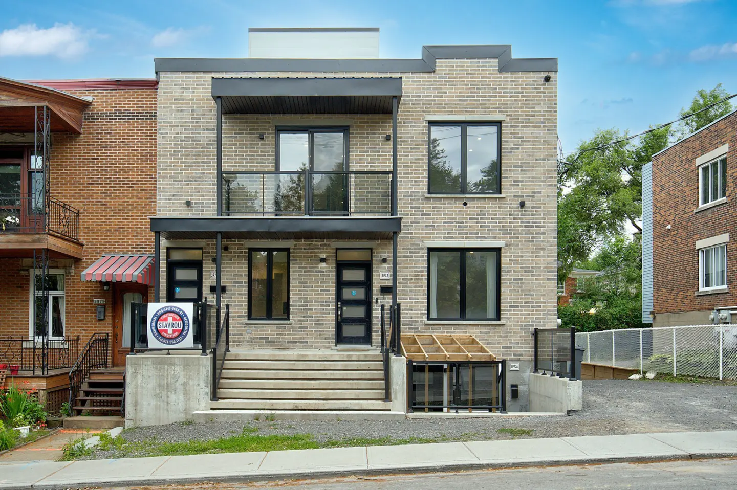 Two-story brick duplex with black trim, balconies, and concrete steps leading to the front doors. A real estate sign is visible.