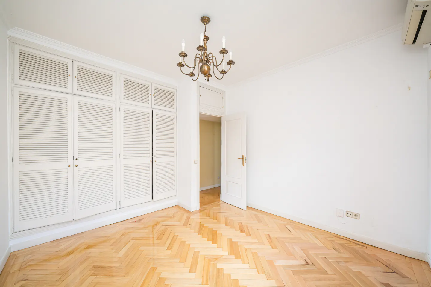 Bright, empty room with herringbone wood floor, white walls, and a white shuttered closet. A brass chandelier hangs from the ceiling.
