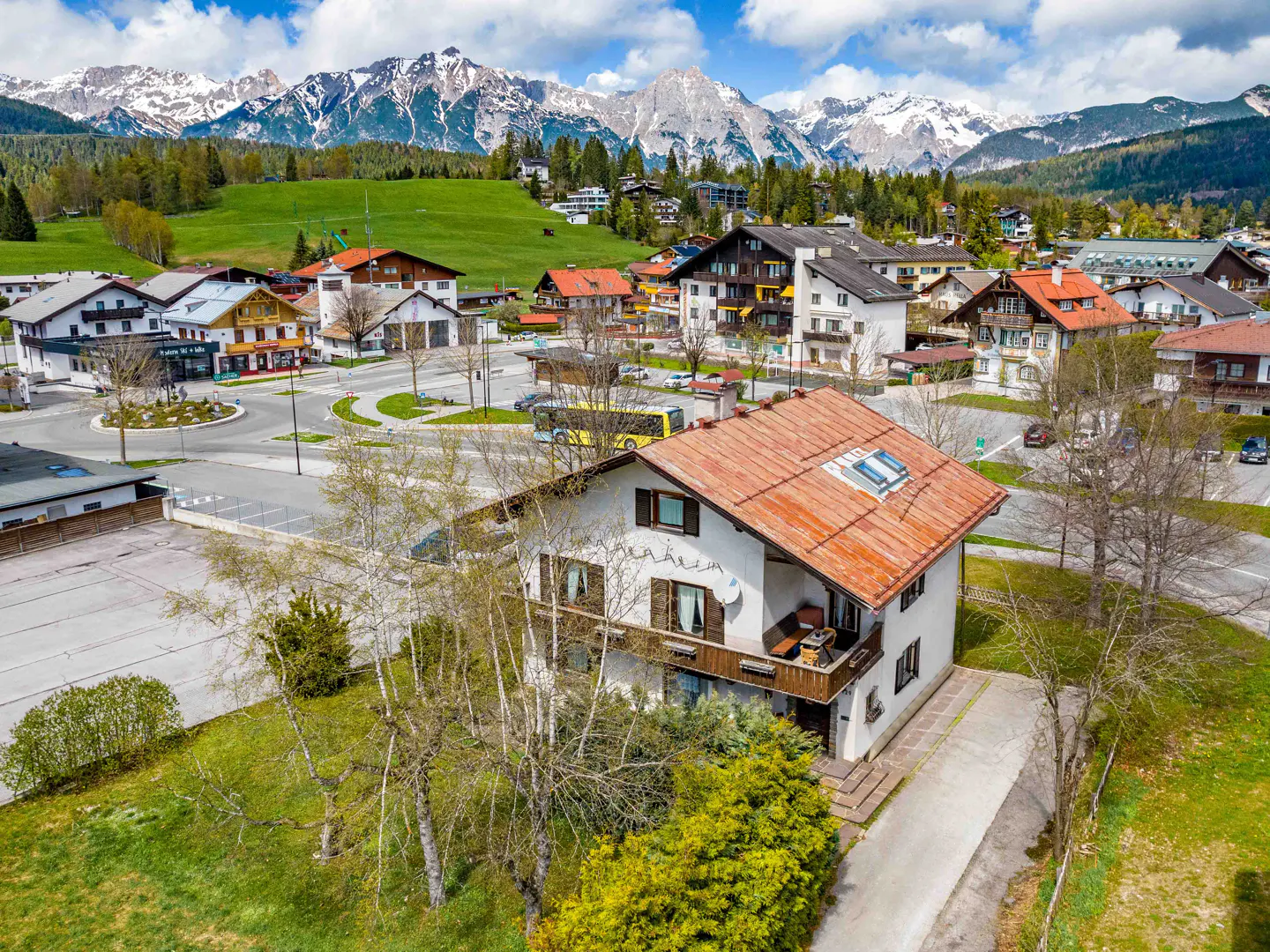 Aerial view of a white house with a red tile roof in a village, mountains in the background.