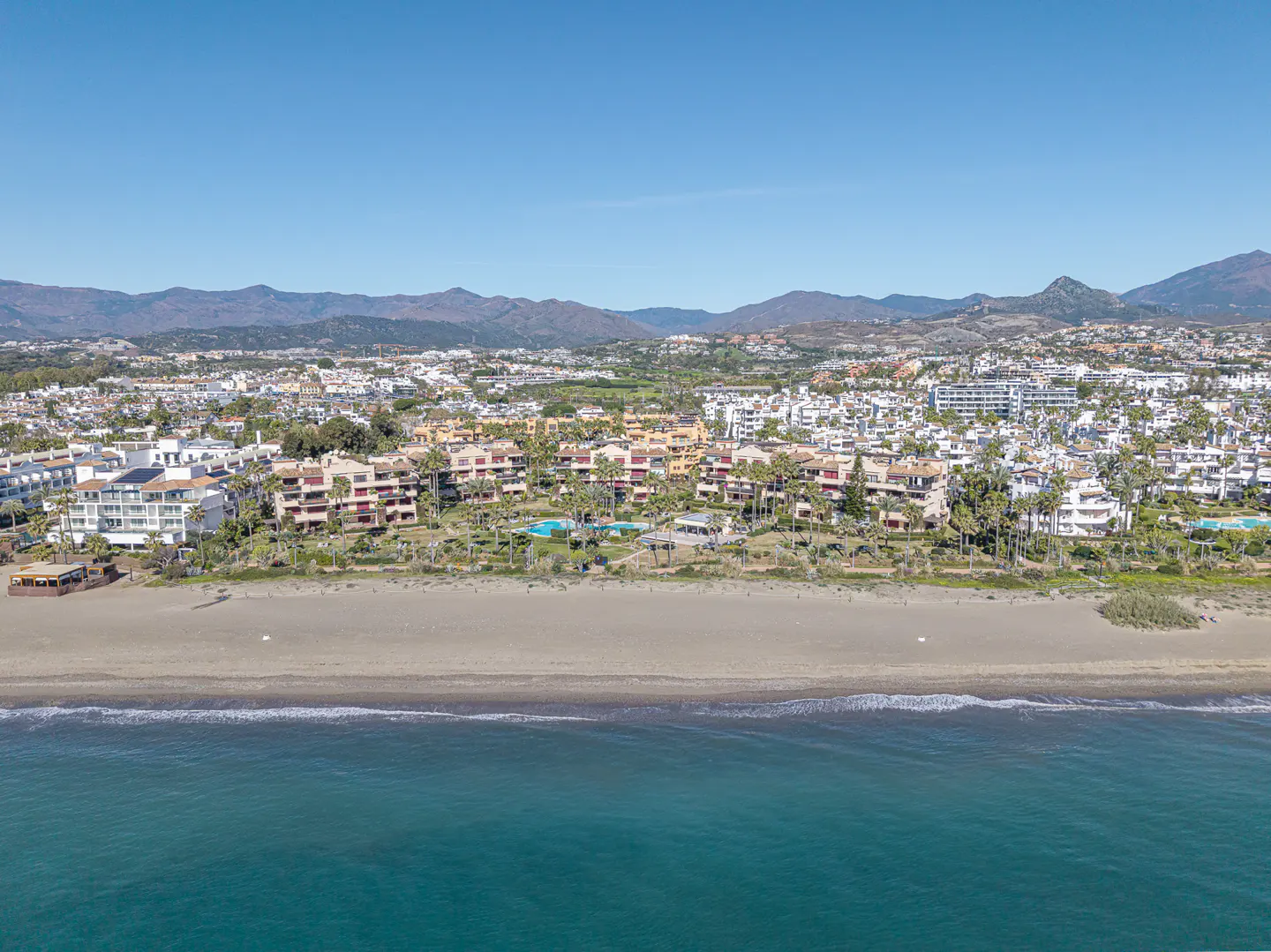 Beachfront property view. Tan buildings line the beach, with palm trees and pools. Blue ocean in the foreground, mountains in the background.
