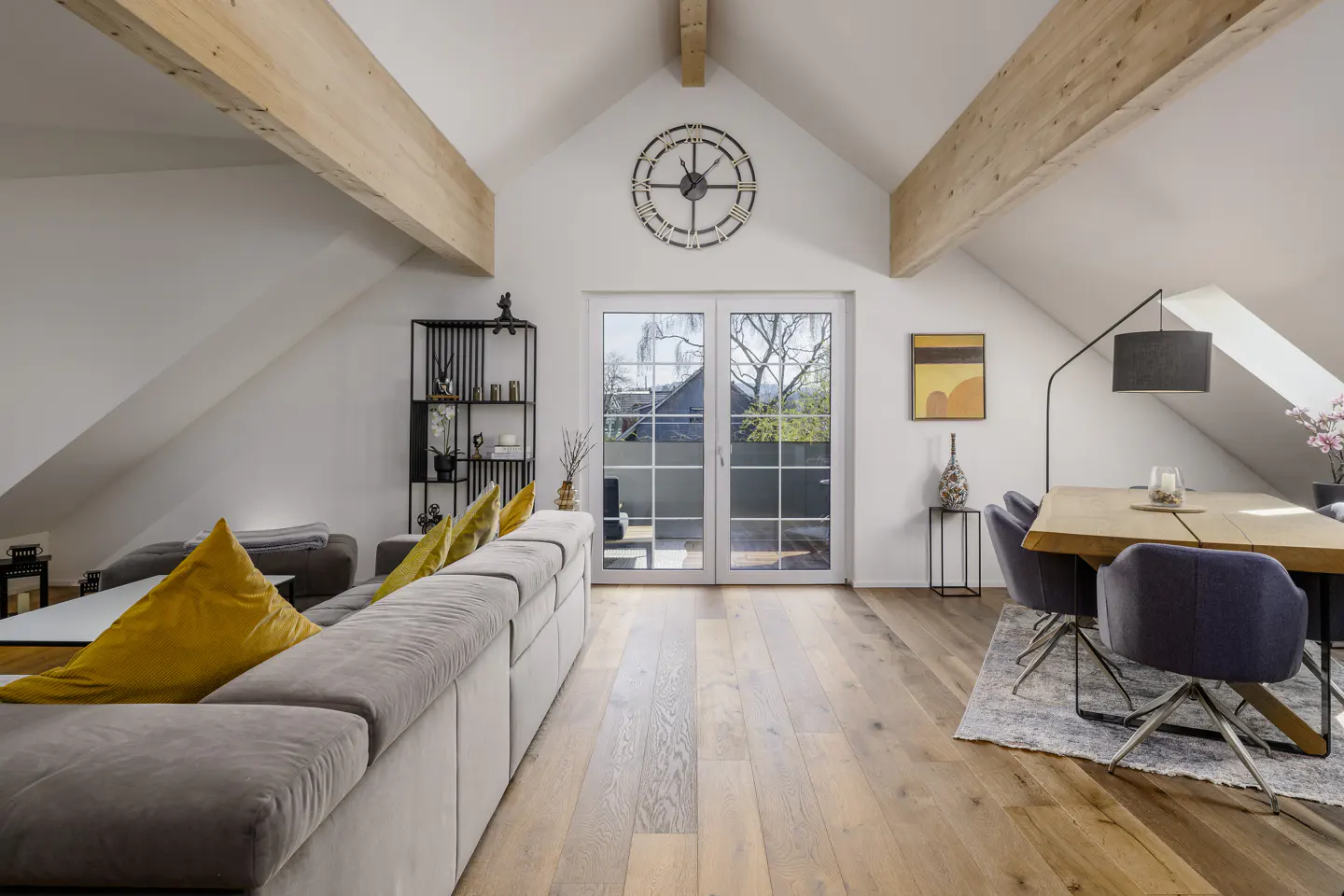 Attic living room with wood floors, exposed beams, and white walls. A gray sofa with yellow pillows faces French doors. Dining table and chairs are on the right.
