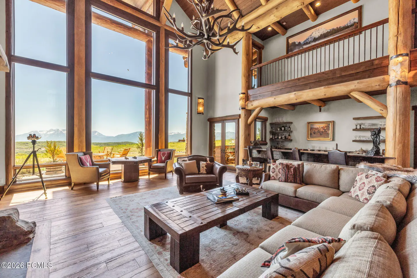A spacious living room with log beams, large windows overlooking mountains, and a rustic chandelier.