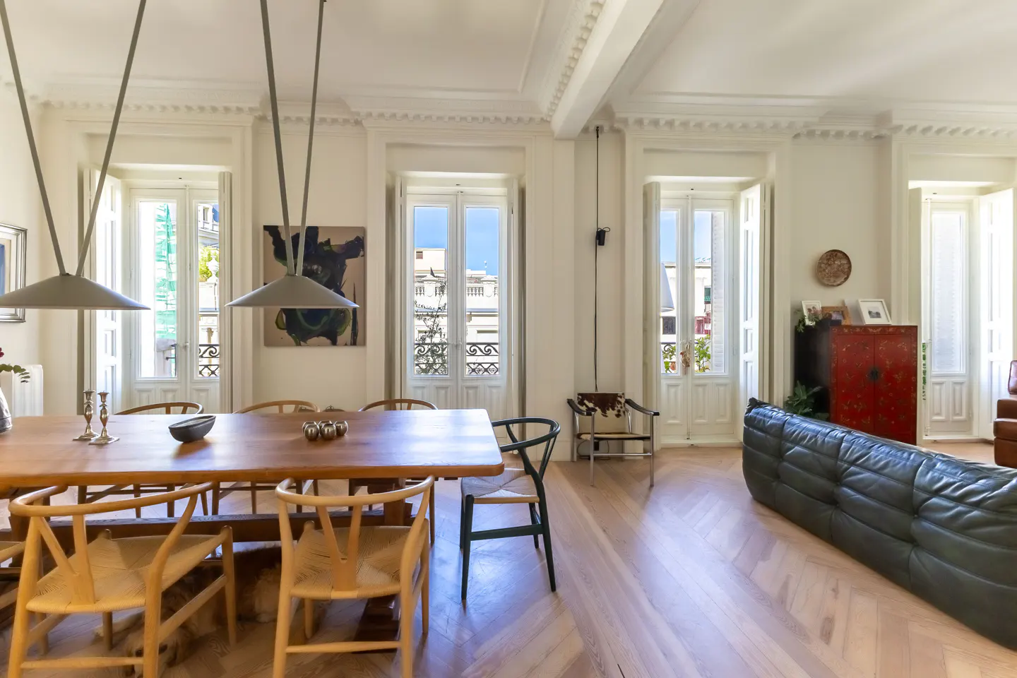 Bright, airy living space with a long wooden table, woven chairs, and a dark green leather sofa. Large windows offer city views.