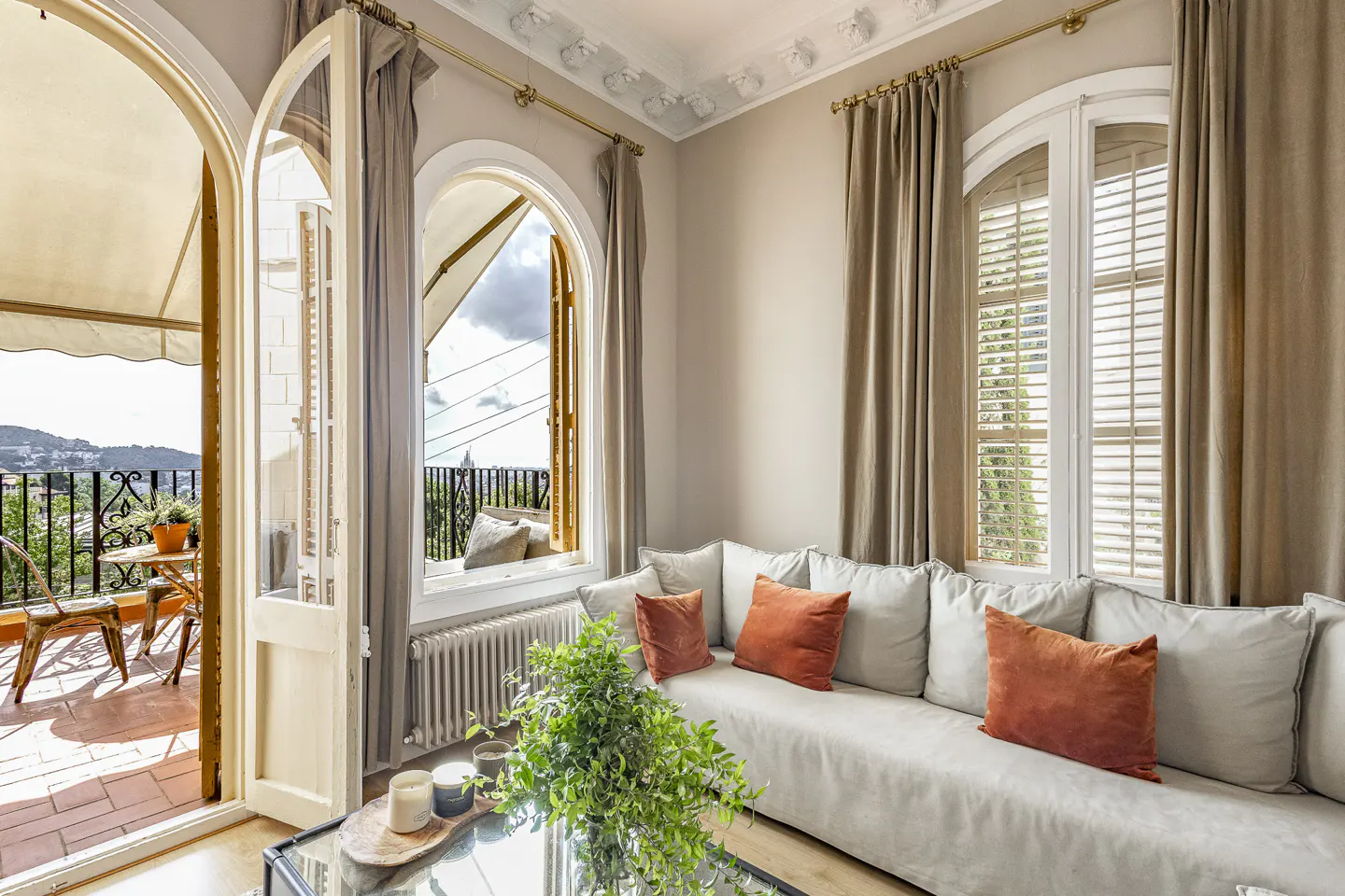Bright living room with a beige sofa, orange cushions, and arched windows opening to a balcony with outdoor seating.