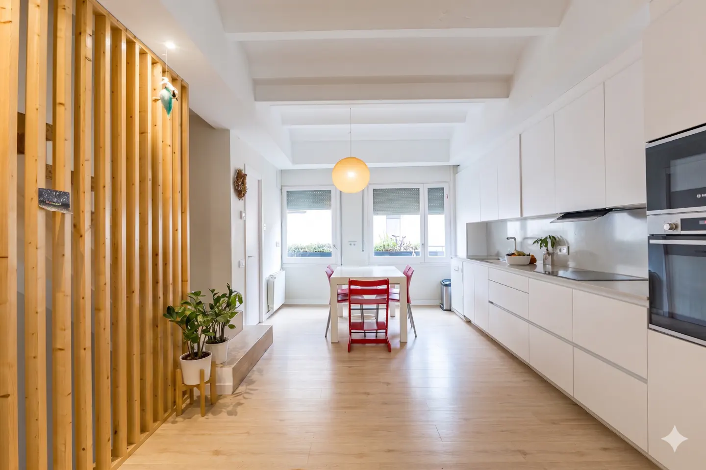 Bright, modern kitchen with light wood floors, white cabinets, and a red dining set near the window. A wooden slat wall adds texture.