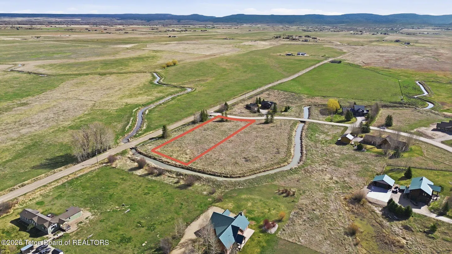 Aerial view of a vacant lot outlined in red, surrounded by green fields, streams, and scattered houses. Mountains are visible in the distance.