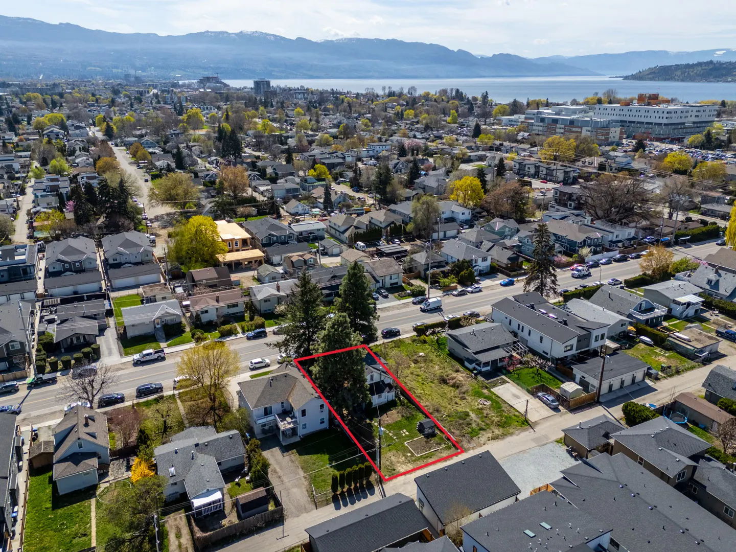 Aerial view of a property outlined in red, surrounded by houses, trees, and a road with cars. A lake and mountains are visible in the background.