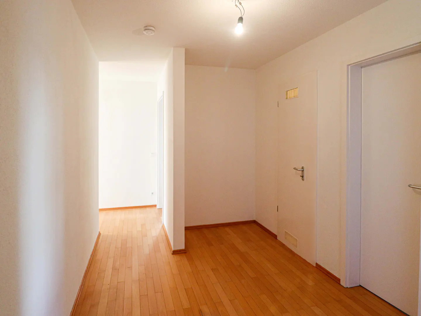 Bright hallway with light wood floors and white walls. Two white doors are visible, one at the end of the hall.