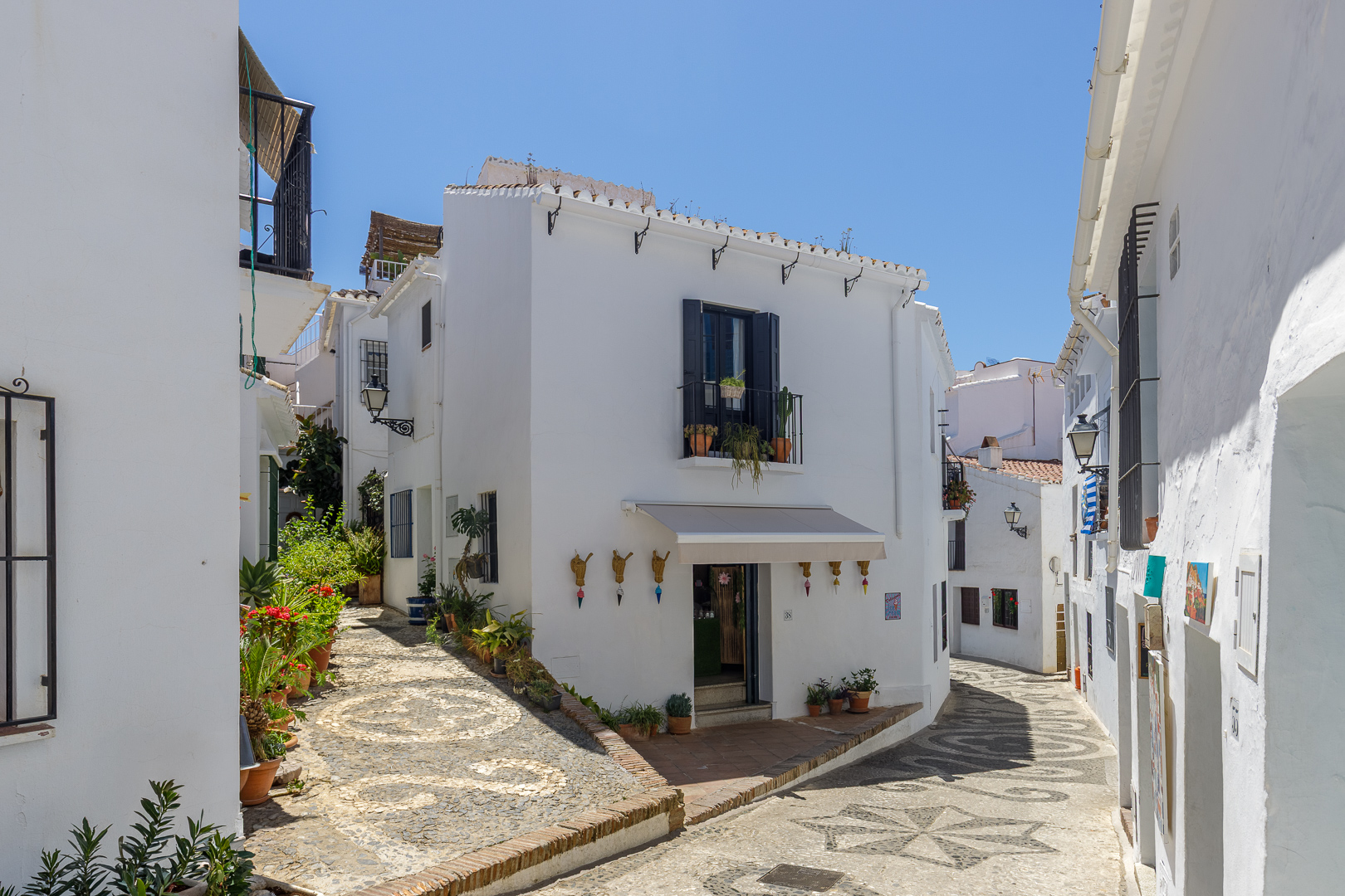 Narrow cobblestone street in a Spanish village with white buildings, potted plants, and decorative wall ornaments.