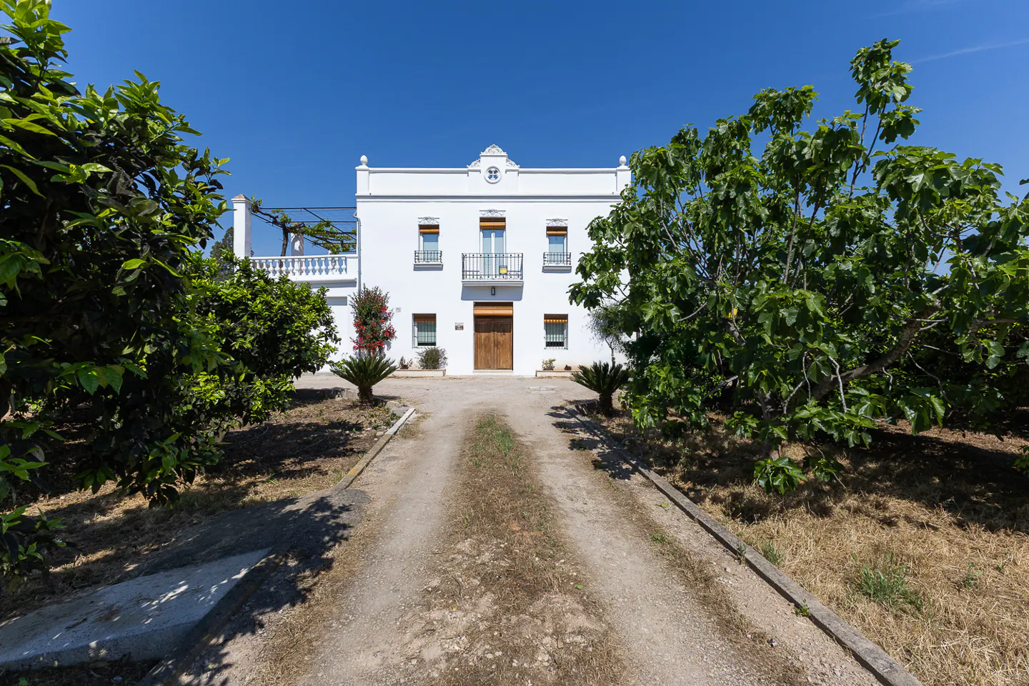 Exterior of a two-story white house with a dirt driveway and green trees under a blue sky.