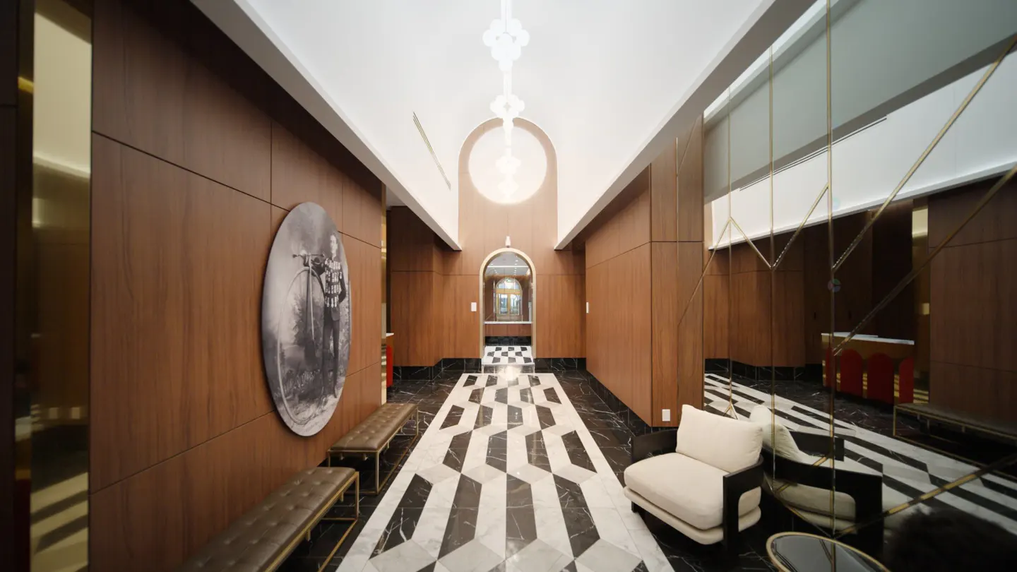Hallway with wood paneled walls, black and white patterned floor, benches, and a white chair. A large mirror is on the right wall.
