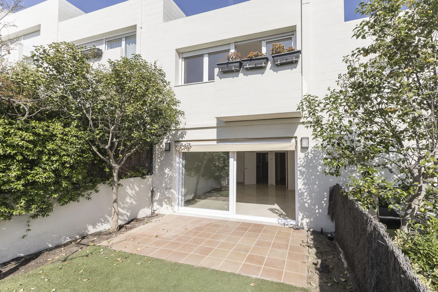 Backyard of a white townhouse with a tiled patio, artificial grass, and trees. Sliding glass doors lead into the house. Flower boxes adorn the window above.