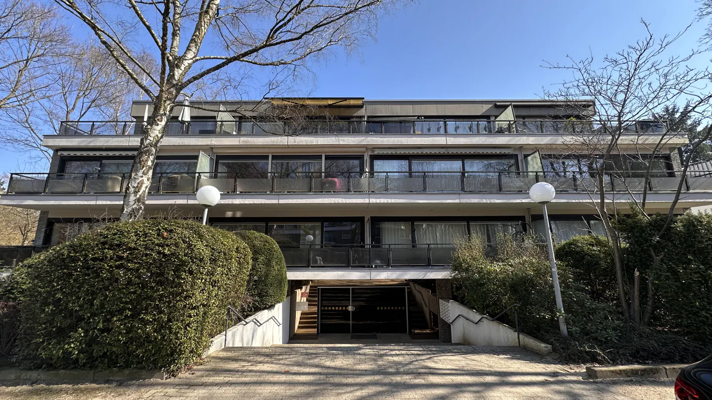 Three-story apartment building with balconies, surrounded by green bushes and trees under a blue sky.