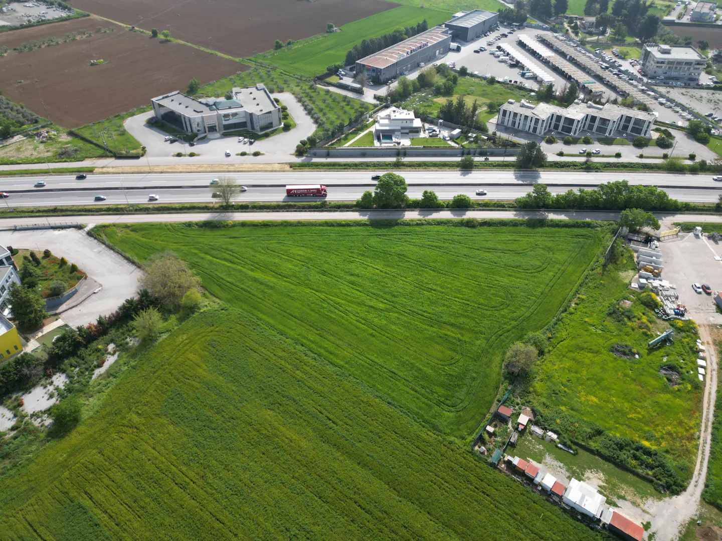 Aerial view of a green field next to a highway with buildings and cars in the background.