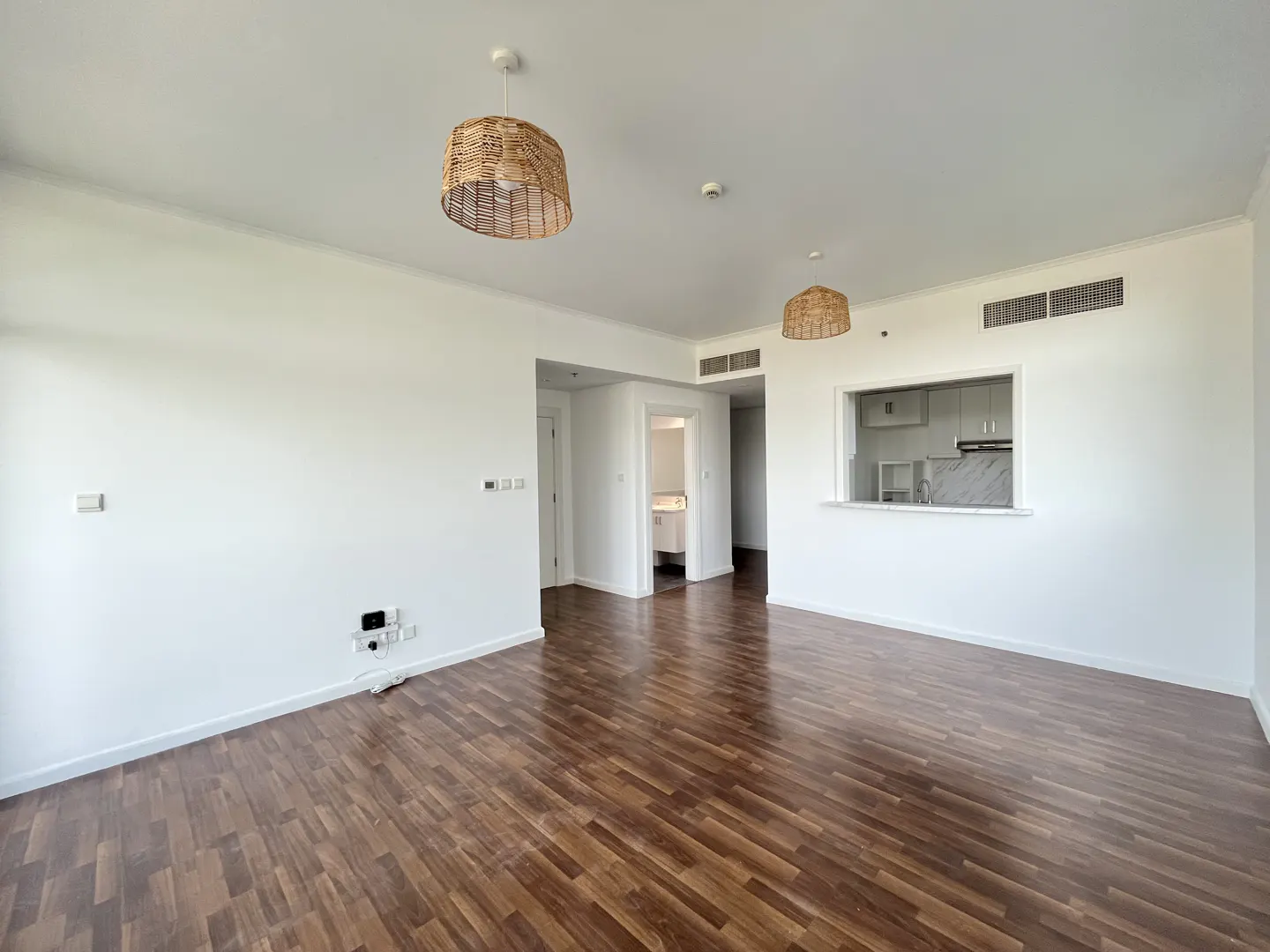 Bright, empty room with wood floors, white walls, and woven pendant lights. A kitchen and bathroom are visible through openings.