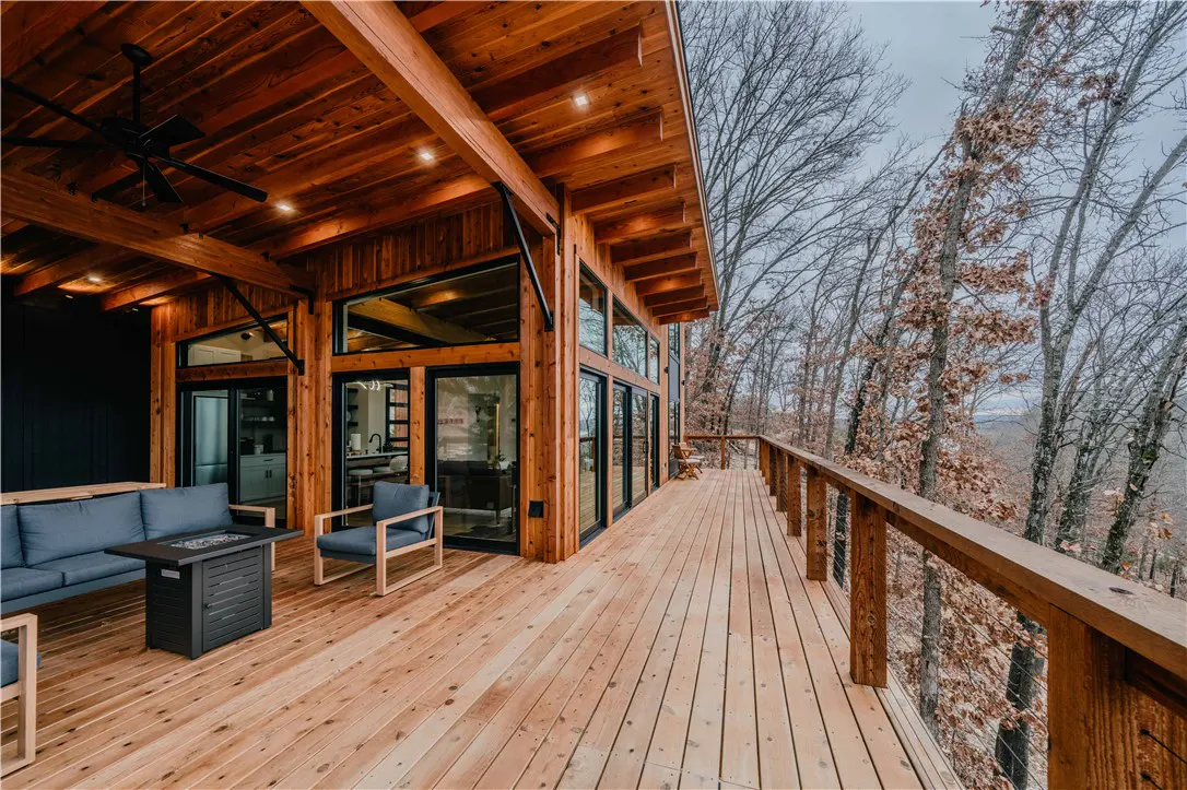 Exterior view of a wood cabin with a deck overlooking a forest. The deck has a sofa, chairs, and a fire pit.