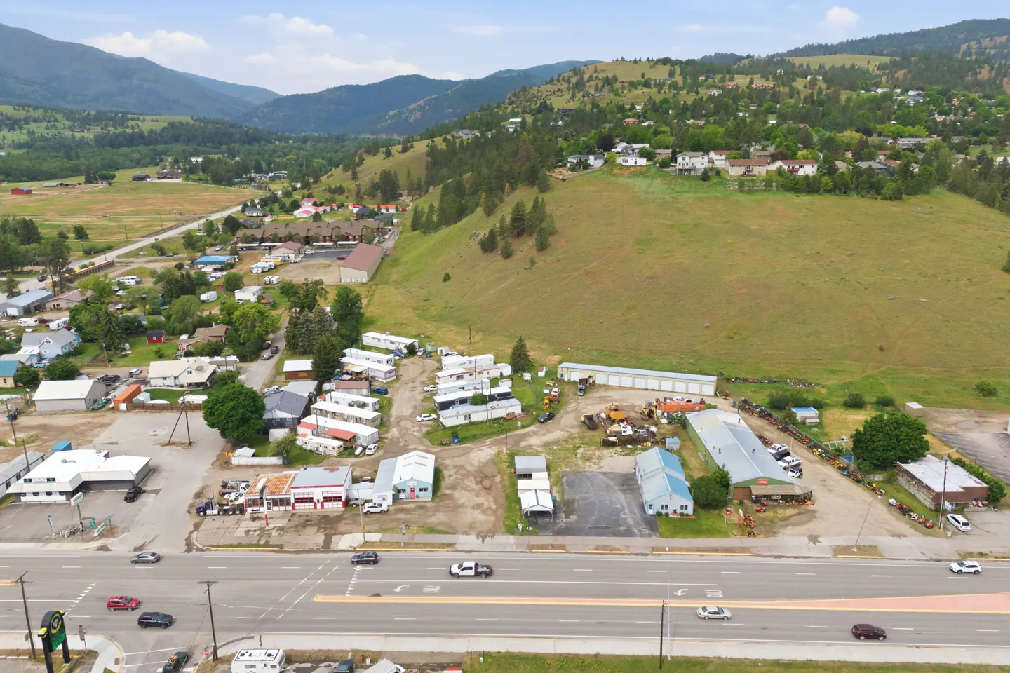 Aerial view of a small town with a highway, commercial buildings, and a mobile home park nestled against grassy hills.