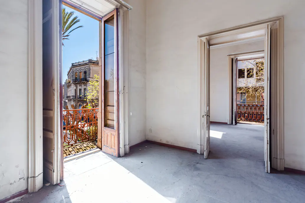 Empty room with open doors leading to balconies with ornate railings and city views. Walls are white, and the floor is gray.
