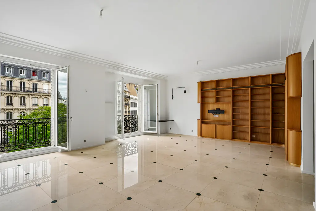 Bright, empty room with light tile floors, a large wooden bookshelf, and French doors opening to a balcony with city views.