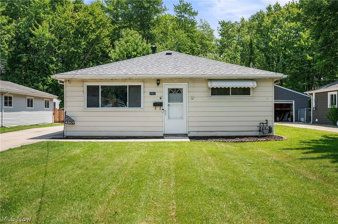 Single-story beige house with a gray shingled roof, white front door, and a green lawn surrounded by lush trees.