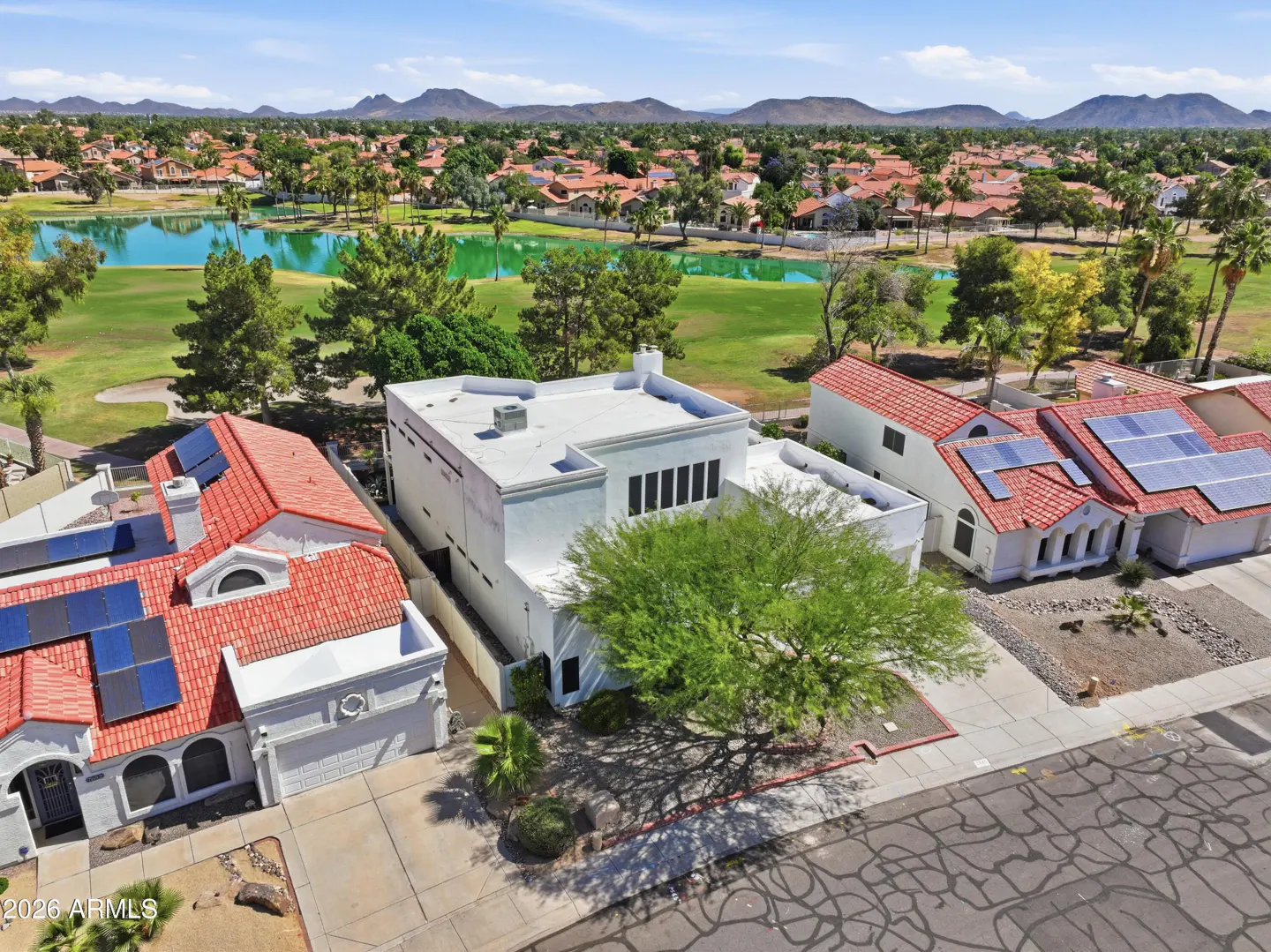 Aerial view of a white, modern house with a flat roof, next to houses with red tile roofs and solar panels, near a green golf course and lake.