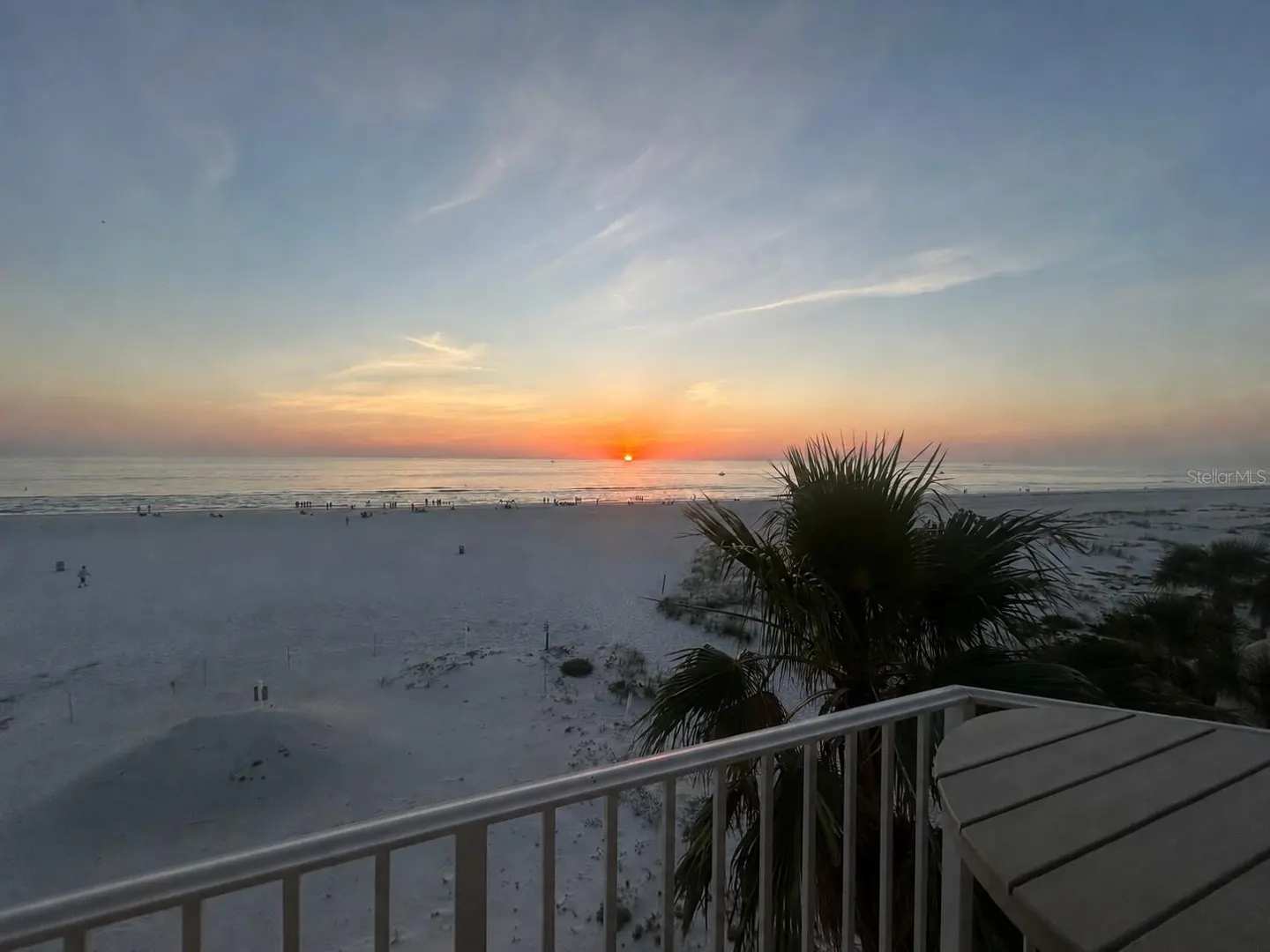 Balcony view of a beach at sunset. The sun is setting over the ocean, casting a warm glow on the sand and water. Palm tree on the right.