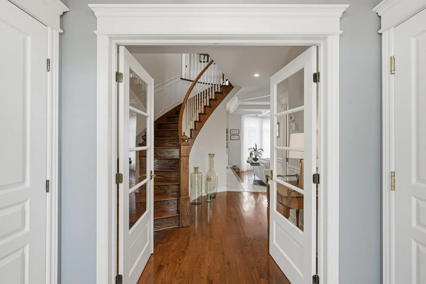 View through white French doors into a foyer with hardwood floors, a curved wooden staircase, and light gray walls.