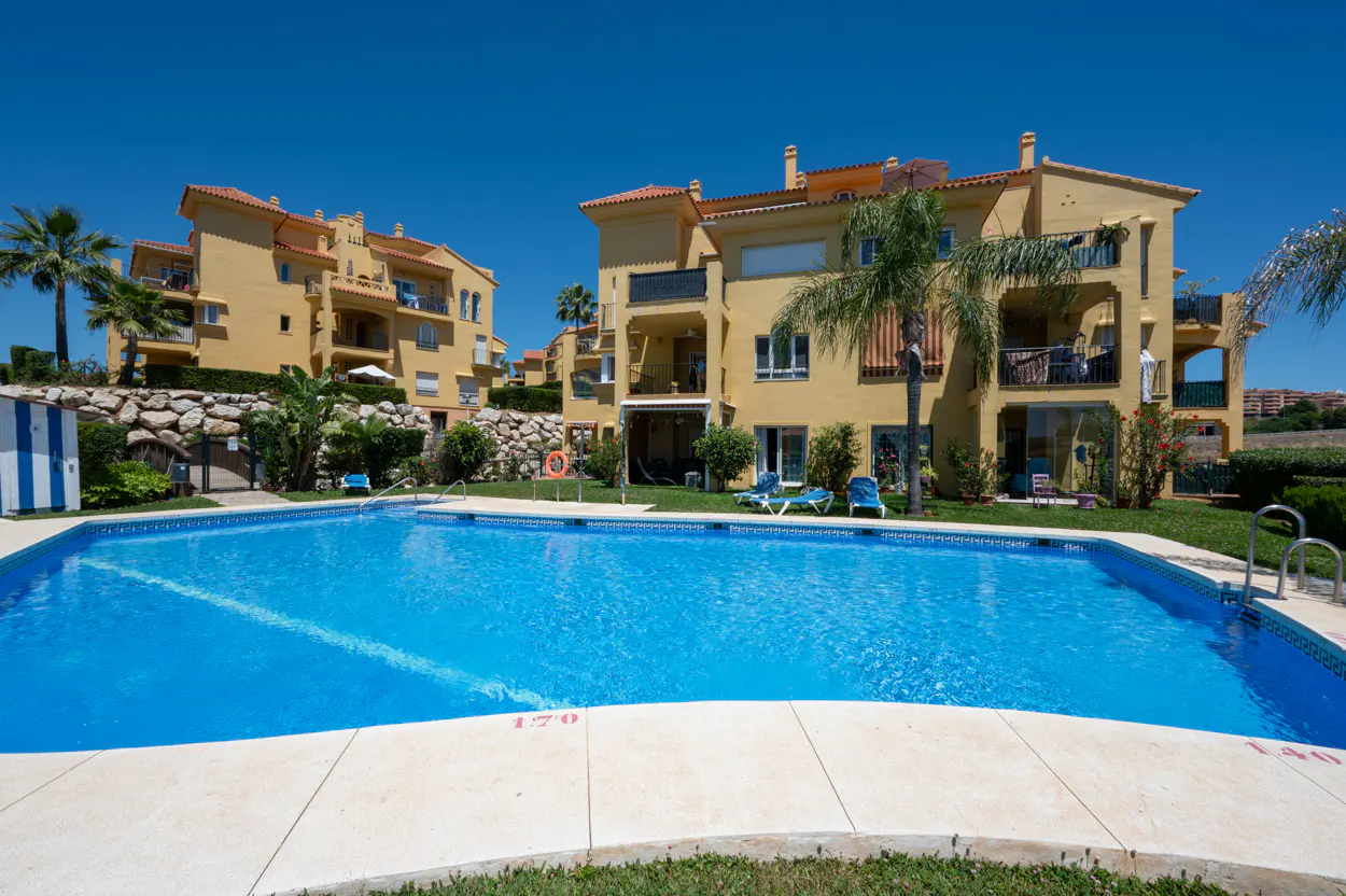 Exterior view of a bright blue swimming pool in front of yellow apartment buildings under a clear blue sky.