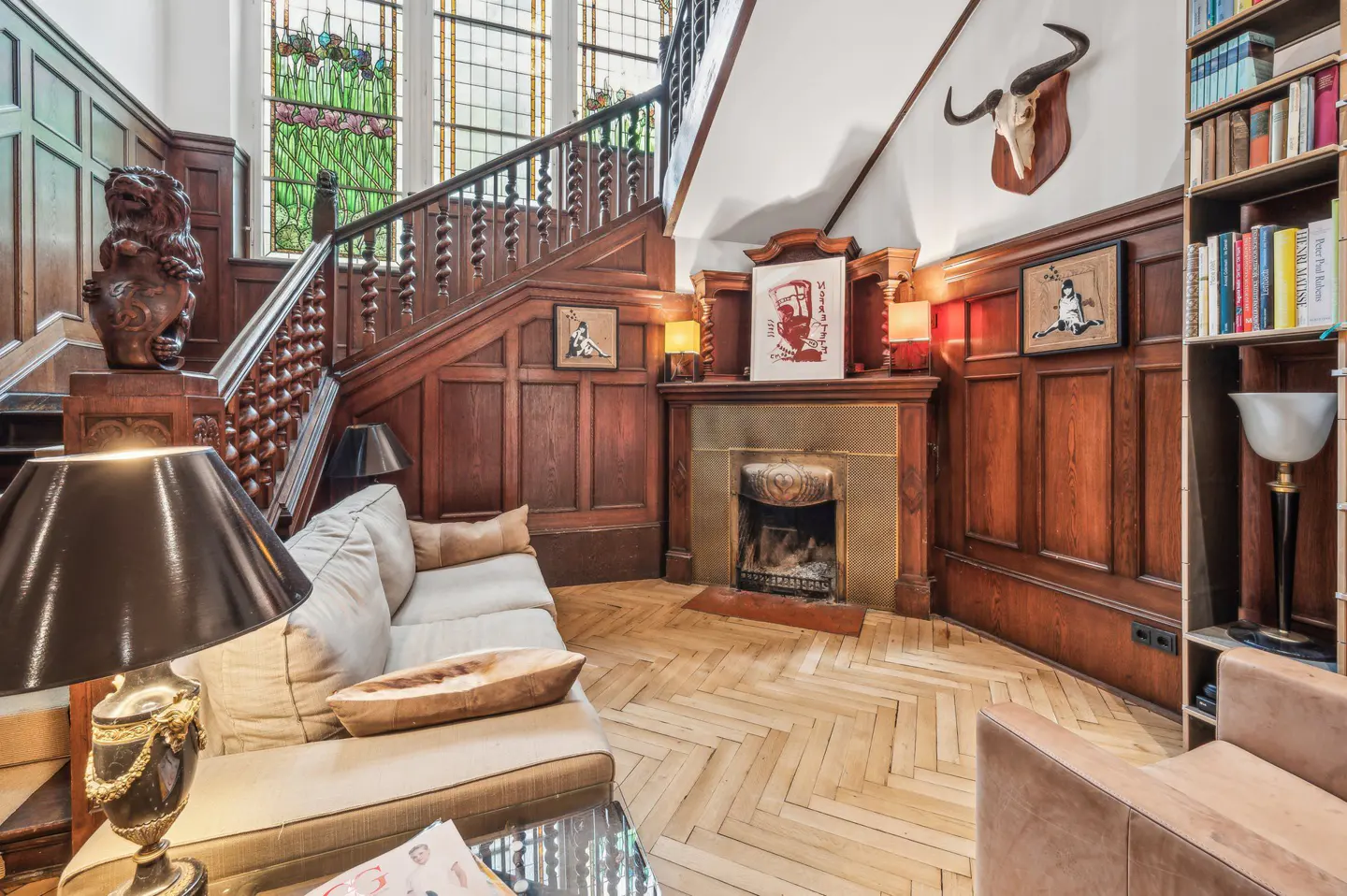 Ornate foyer with wood paneling, herringbone floors, and a staircase with a carved lion. A fireplace, sofa, and bookshelf add to the room's character.