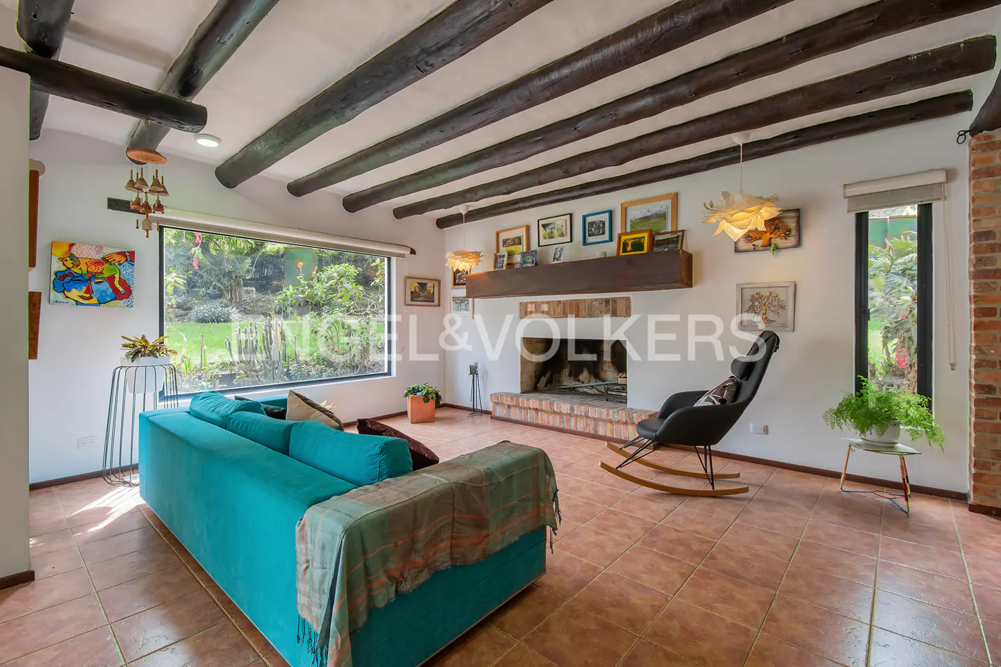 Living room with a teal sofa, fireplace, rocking chair, and exposed wood beams on the ceiling. Large window with a view of the garden.