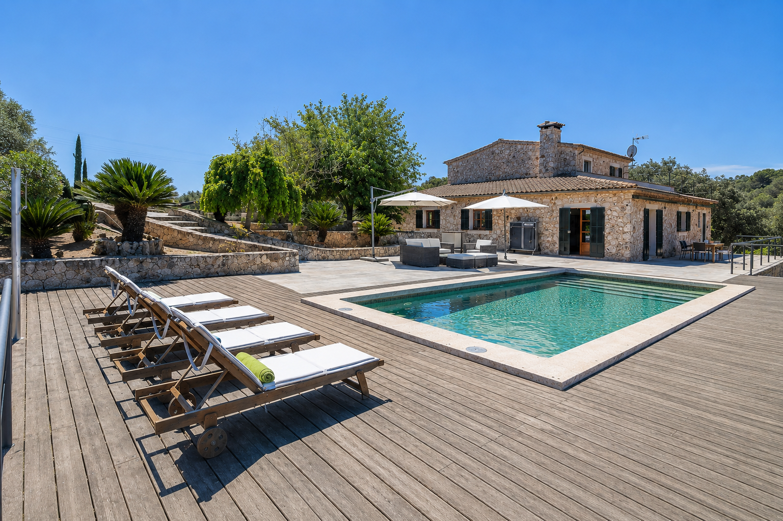 Stone villa with a swimming pool, wooden deck, lounge chairs, and patio furniture under a clear blue sky.