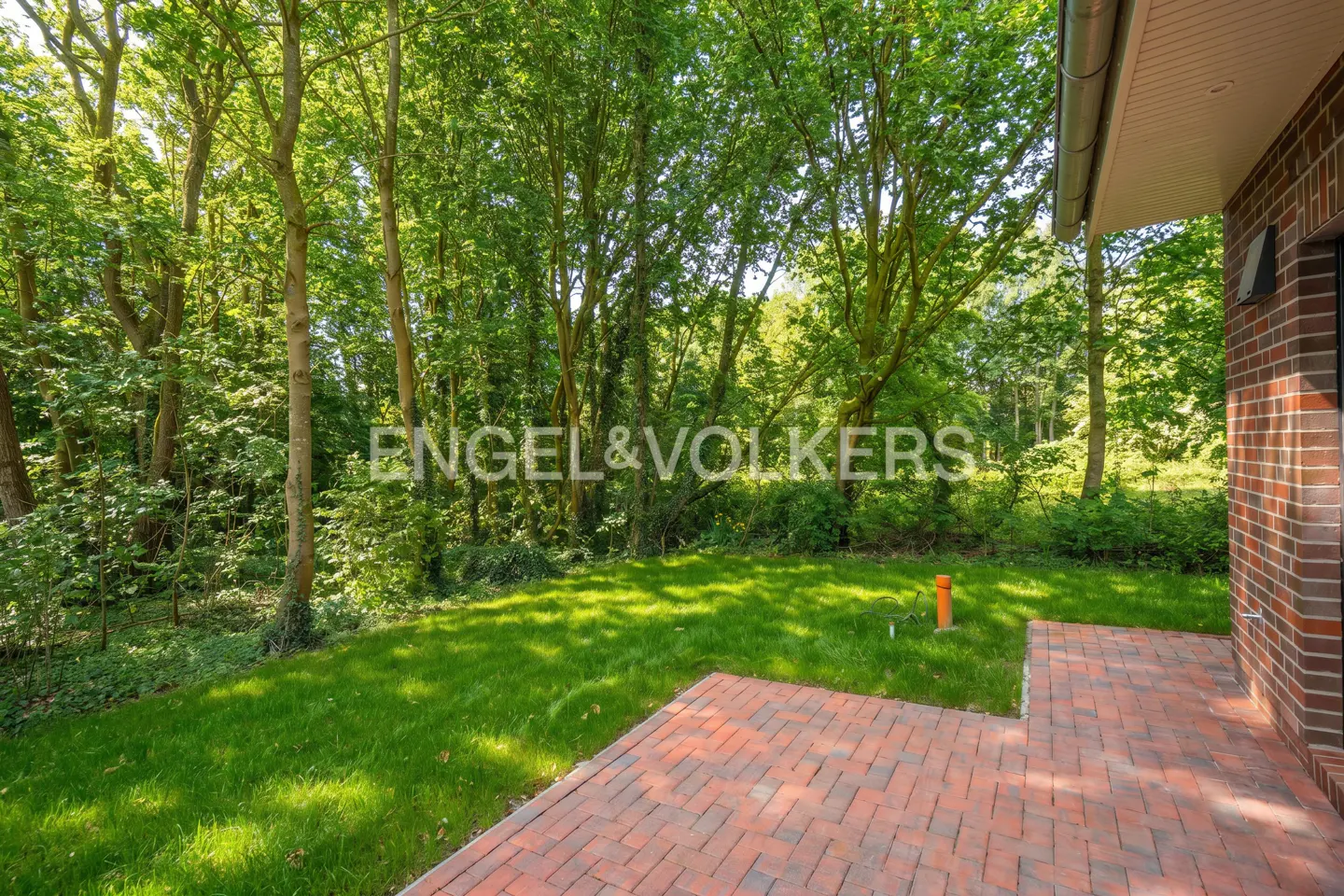 Brick patio and lawn lead to a wooded area. Engel & Volkers logo is superimposed on the lawn.