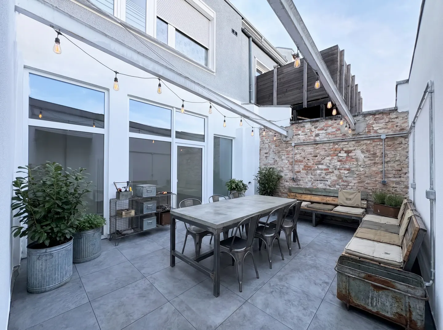 Outdoor patio with a concrete table, metal chairs, and string lights. A brick wall and potted plants add character to the space.