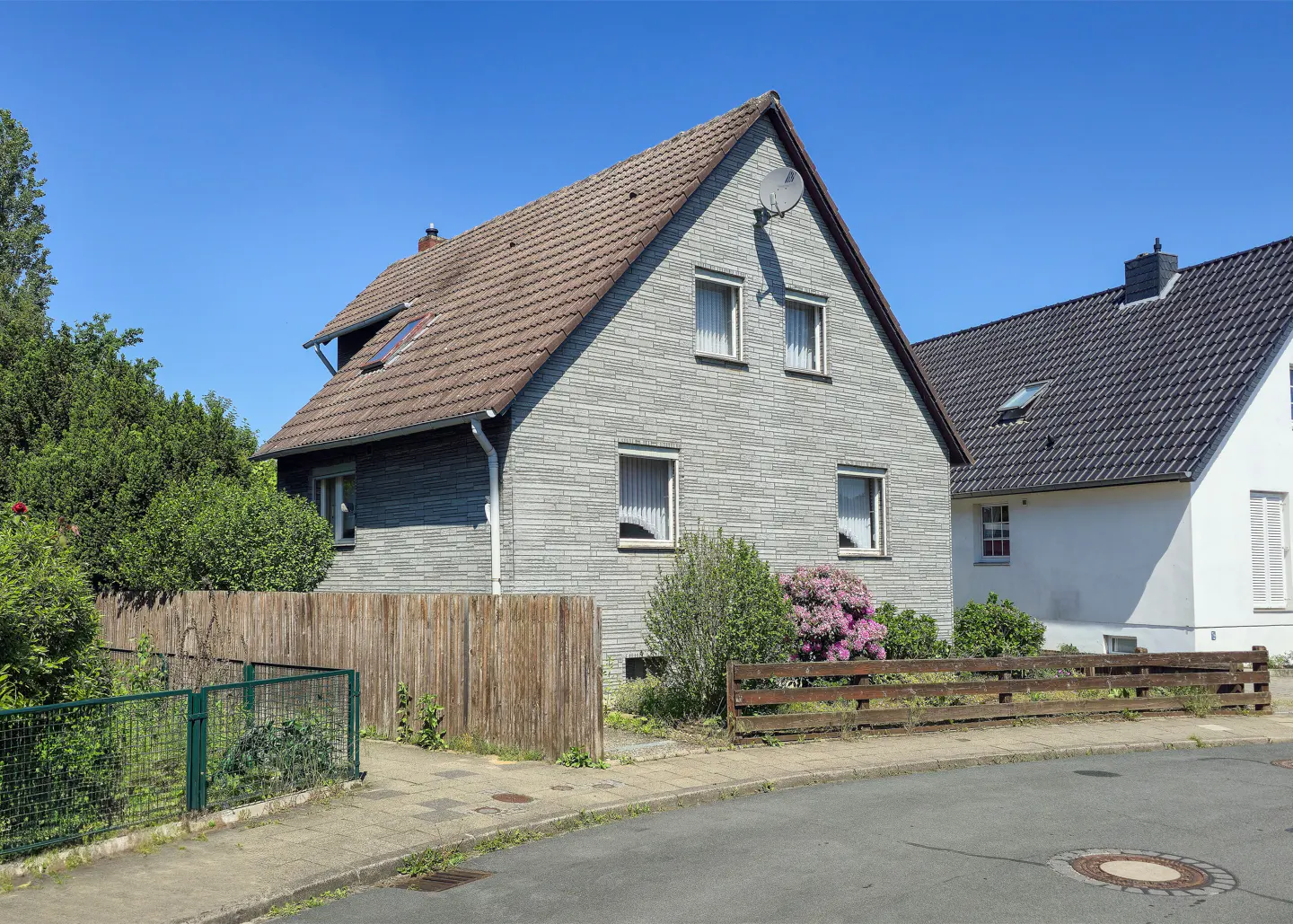 Two-story house with gray brick and brown roof, satellite dish, and white window trim. A wooden fence and green foliage surround the property.