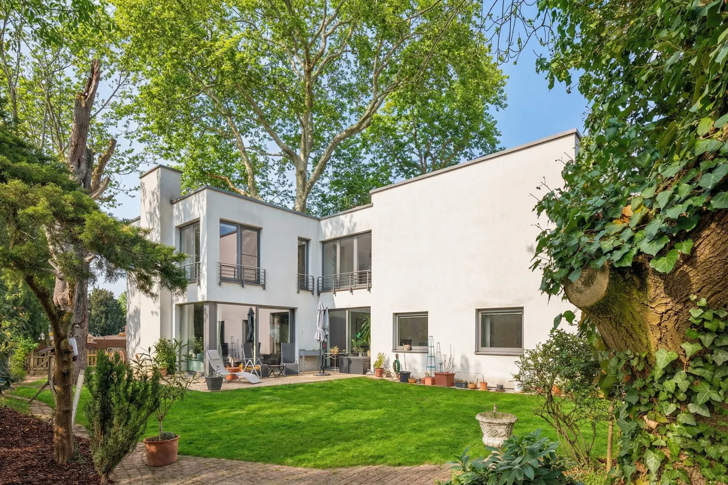 Modern white house with a green lawn and patio furniture, surrounded by trees and greenery.