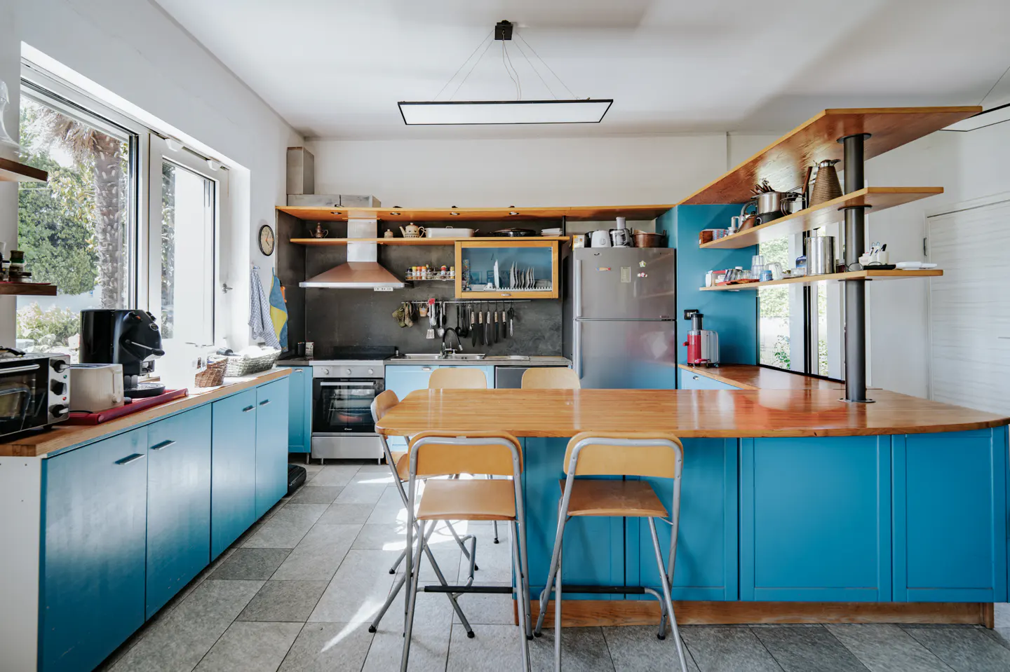A bright kitchen with blue cabinets, a wood countertop island with chairs, and open shelving. Stainless steel appliances and a large window are visible.