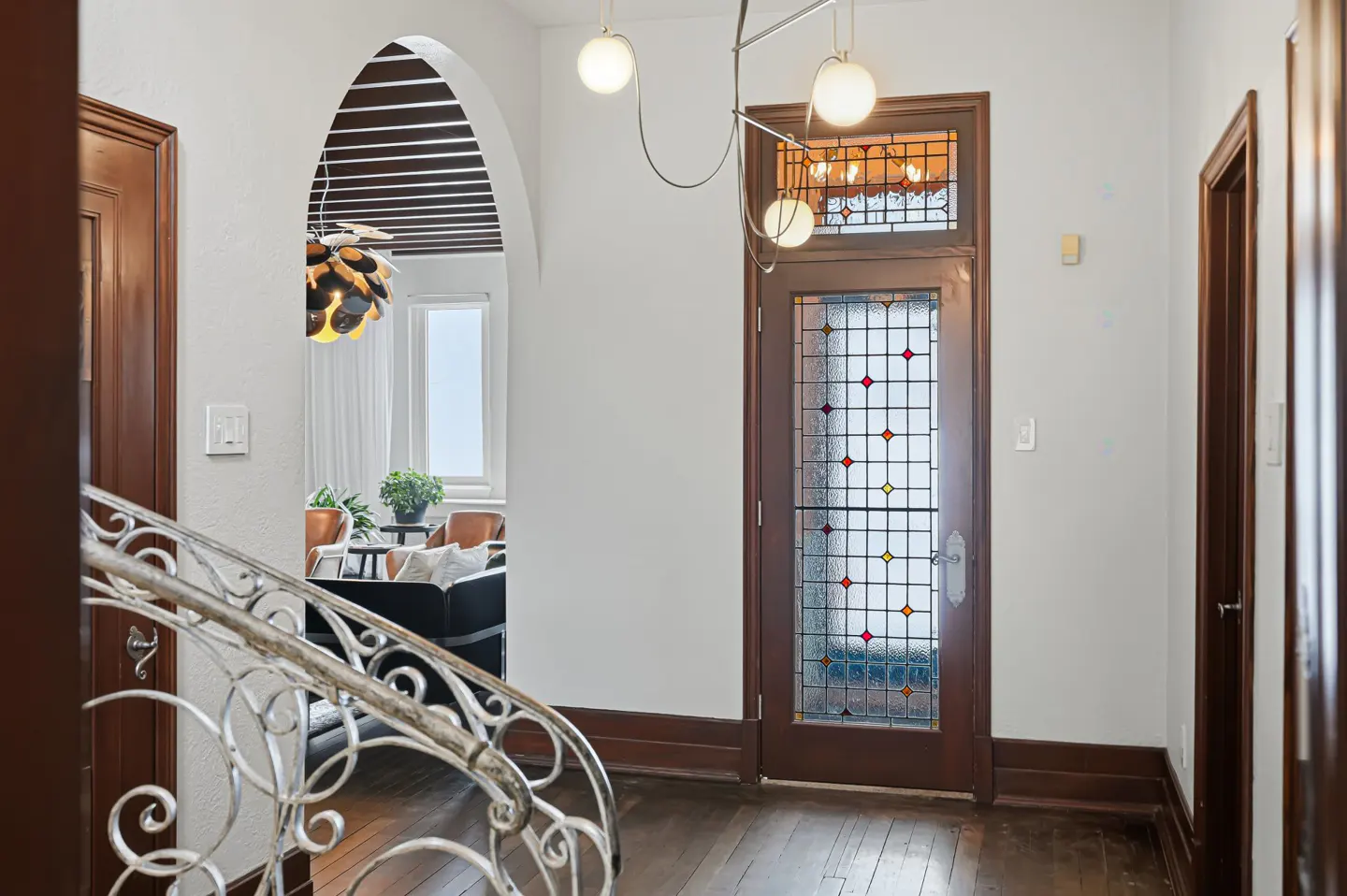 Hallway with a stained glass door, white walls, and dark wood trim. A wrought iron staircase is on the left. A living room is visible through an archway.