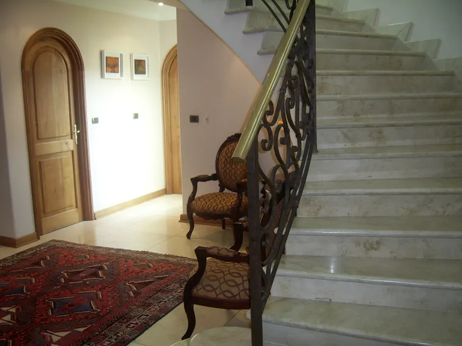 Interior view of a foyer with marble stairs, ornate iron railing, two chairs, a red rug, and arched wooden doors.