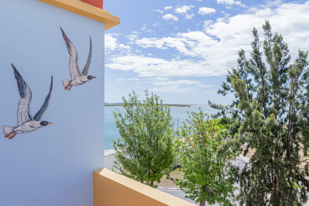 Exterior view of a light blue building with two painted seagulls, overlooking trees and a body of water under a partly cloudy sky.