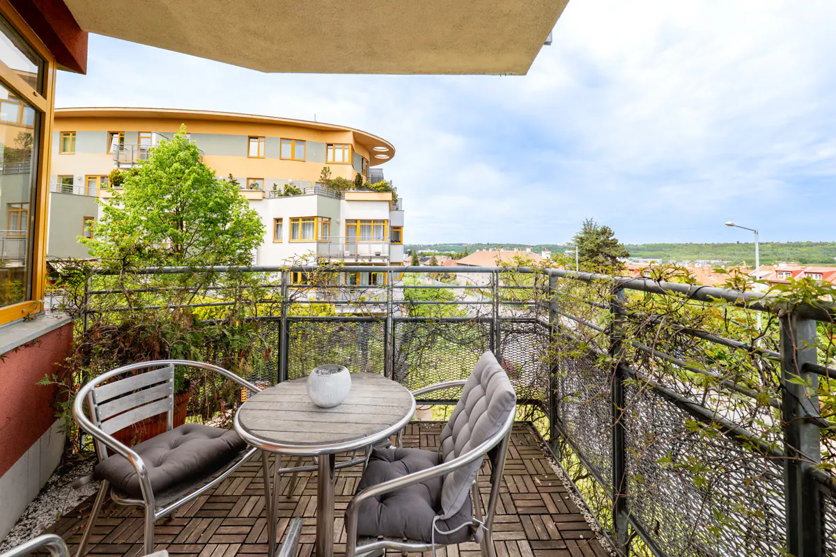 Balcony view with a round table, two chairs with gray cushions, and a metal railing with climbing plants overlooking a cityscape.