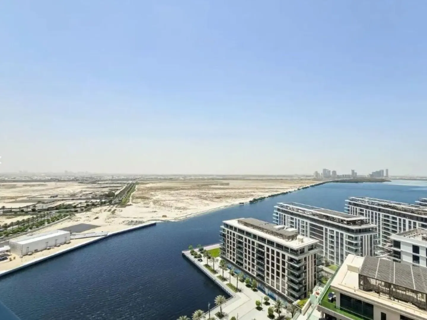 Aerial view of modern white apartment buildings along a blue canal, with desert and city skyline in the background under a clear sky.