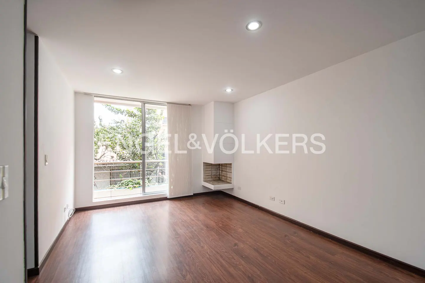 Bright, empty room with hardwood floors, white walls, and a large window overlooking greenery. A modern fireplace is built into the wall.