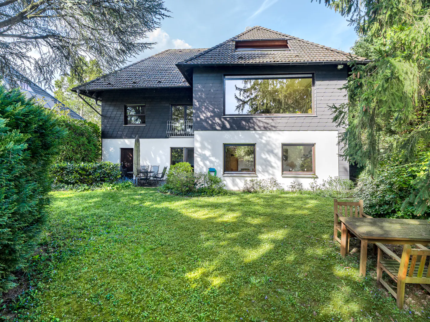 Backyard view of a two-story house with a gray roof, dark siding, and white stucco. Green lawn, trees, and patio furniture are visible.